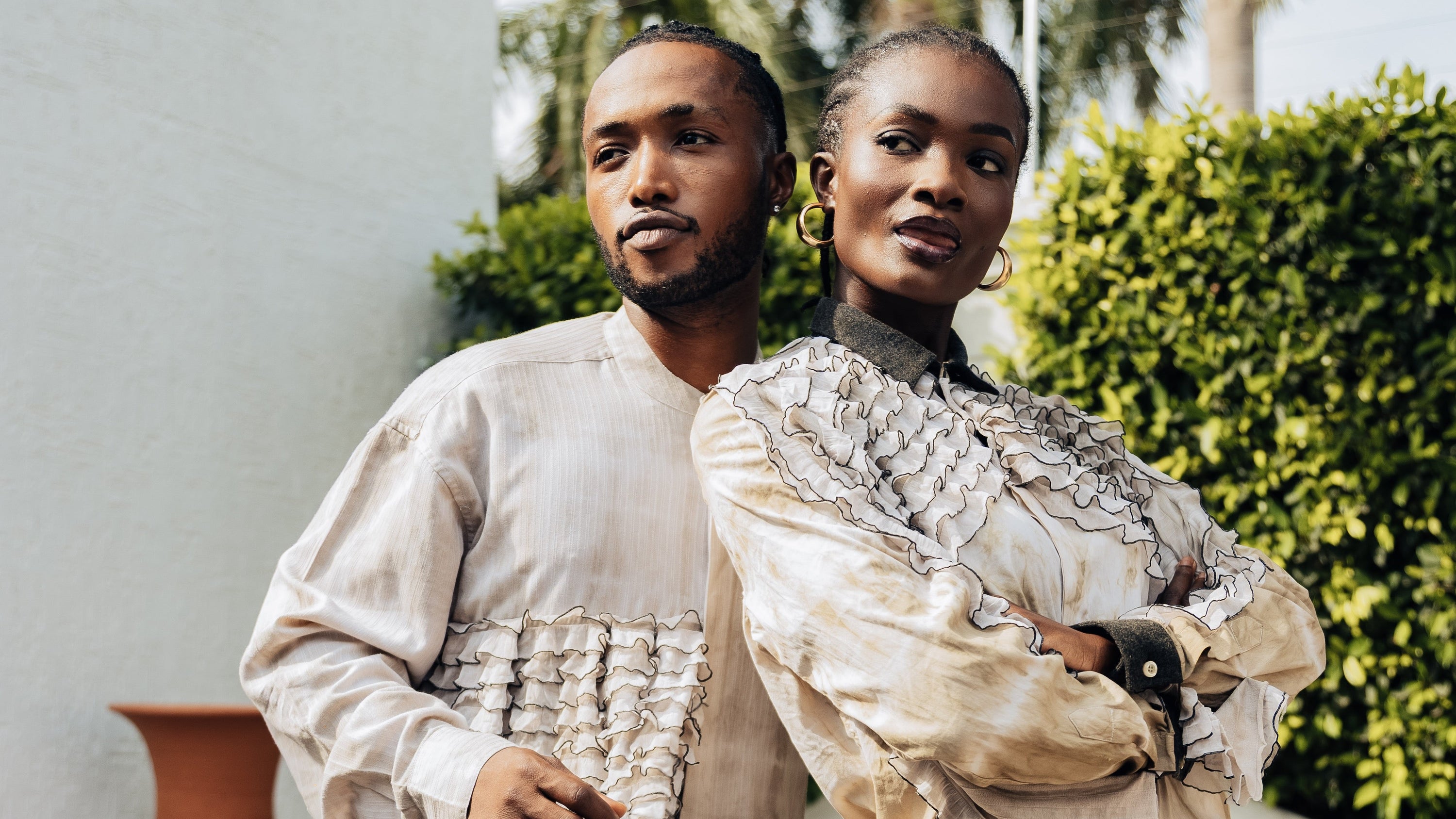 Two people wearing textured, layered outfits standing outdoors with greenery and a white wall in the background.