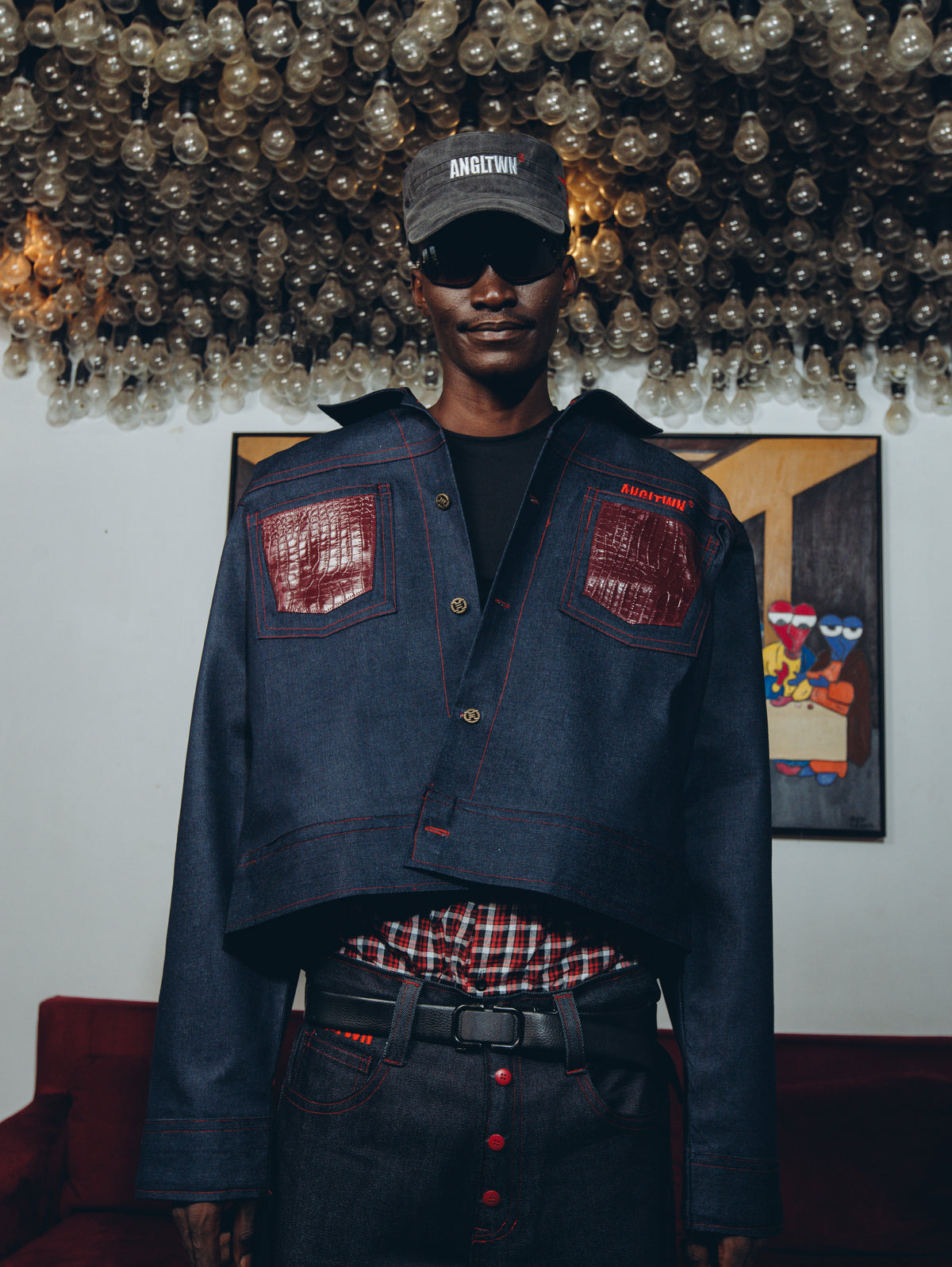 A model wearing a dark blue cropped denim jacket with red contrast stitching and red patch pockets, styled with a red plaid shirt, dark jeans, and a dark gray cap with 'ANTIGONY' printed on it, standing in front of a wall with a light fixture.