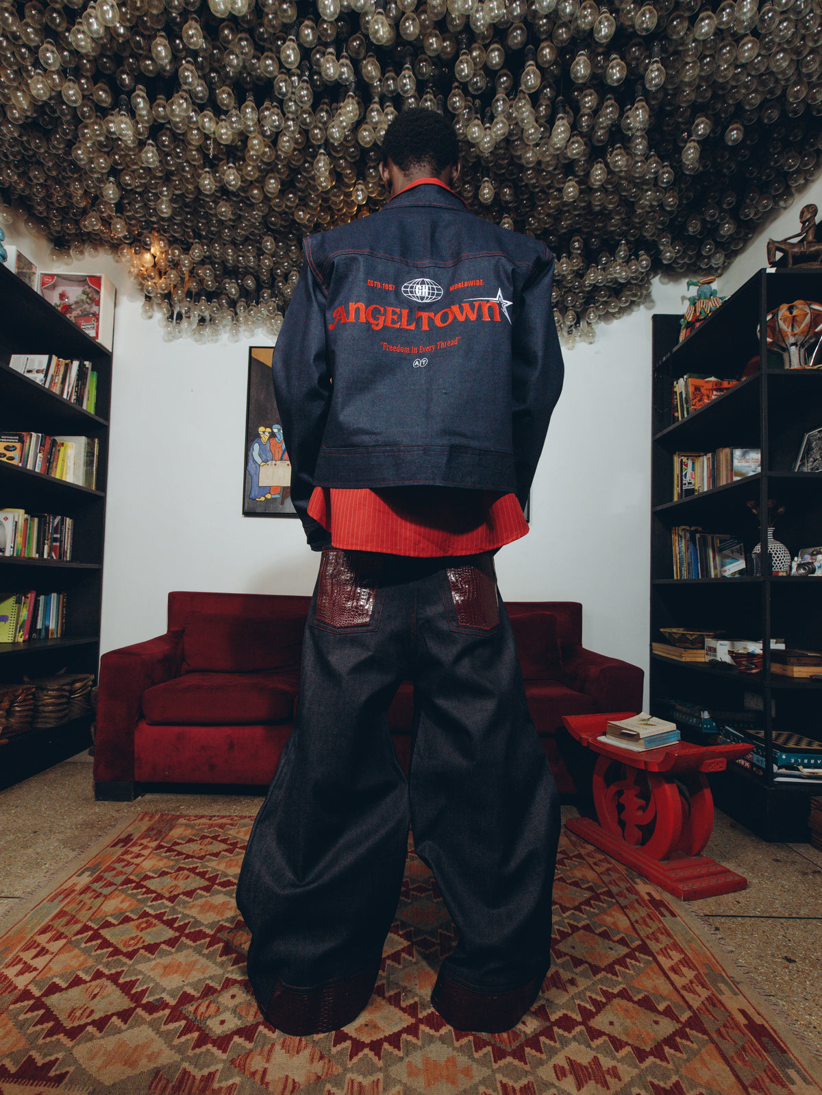 A person stands with their back to the camera wearing a dark denim jacket with 'ANGELTOWN' printed in red across the back, layered over a red shirt and matching dark baggy pants. The setting is a cozy, cluttered room with bookshelves, a red couch, and a patterned rug.