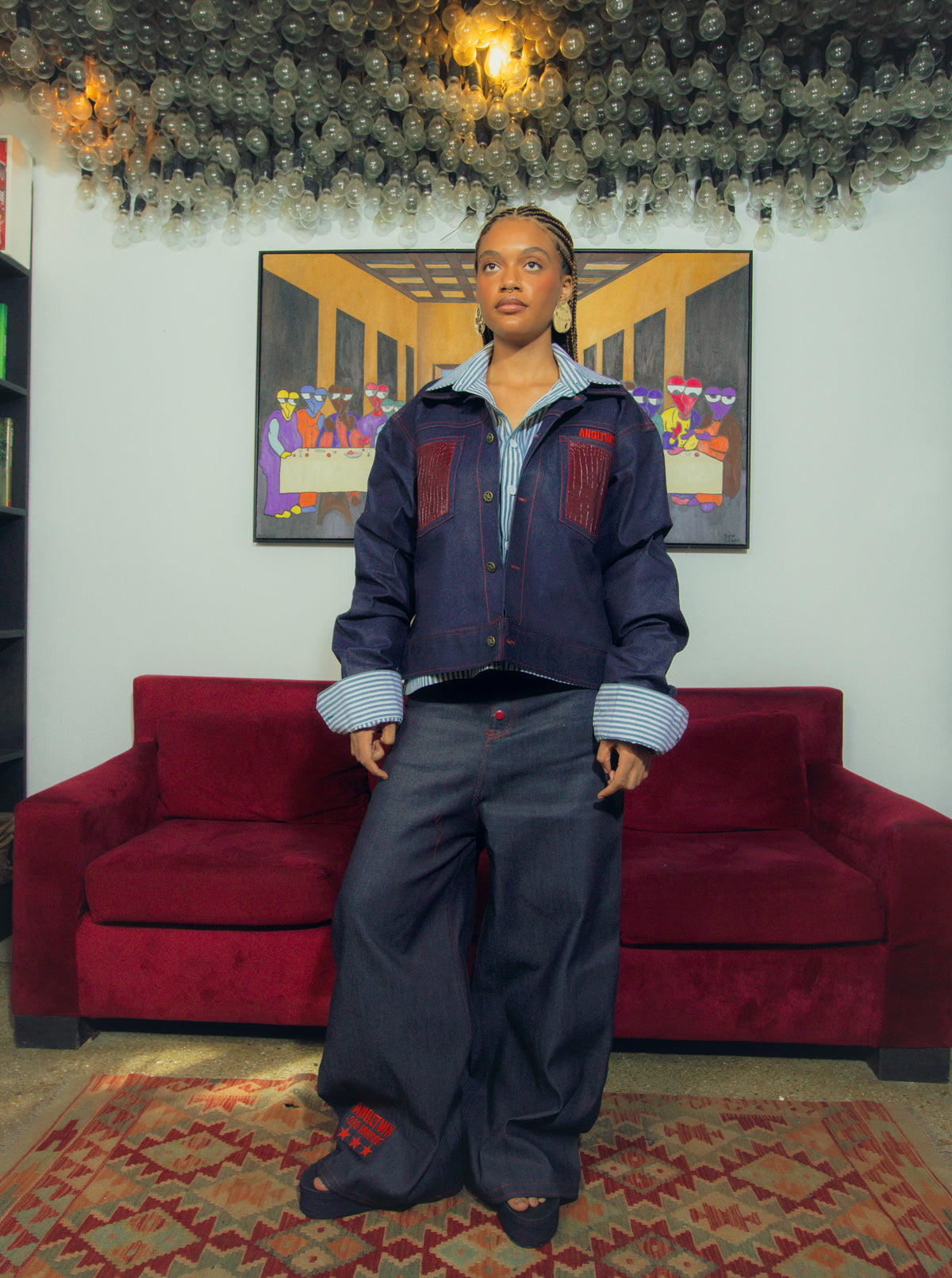 A model stands in a room wearing a dark blue denim jacket with red patch pockets, a light blue striped shirt, and dark wide-leg jeans. Behind her is a red velvet sofa and a framed painting on a white wall.