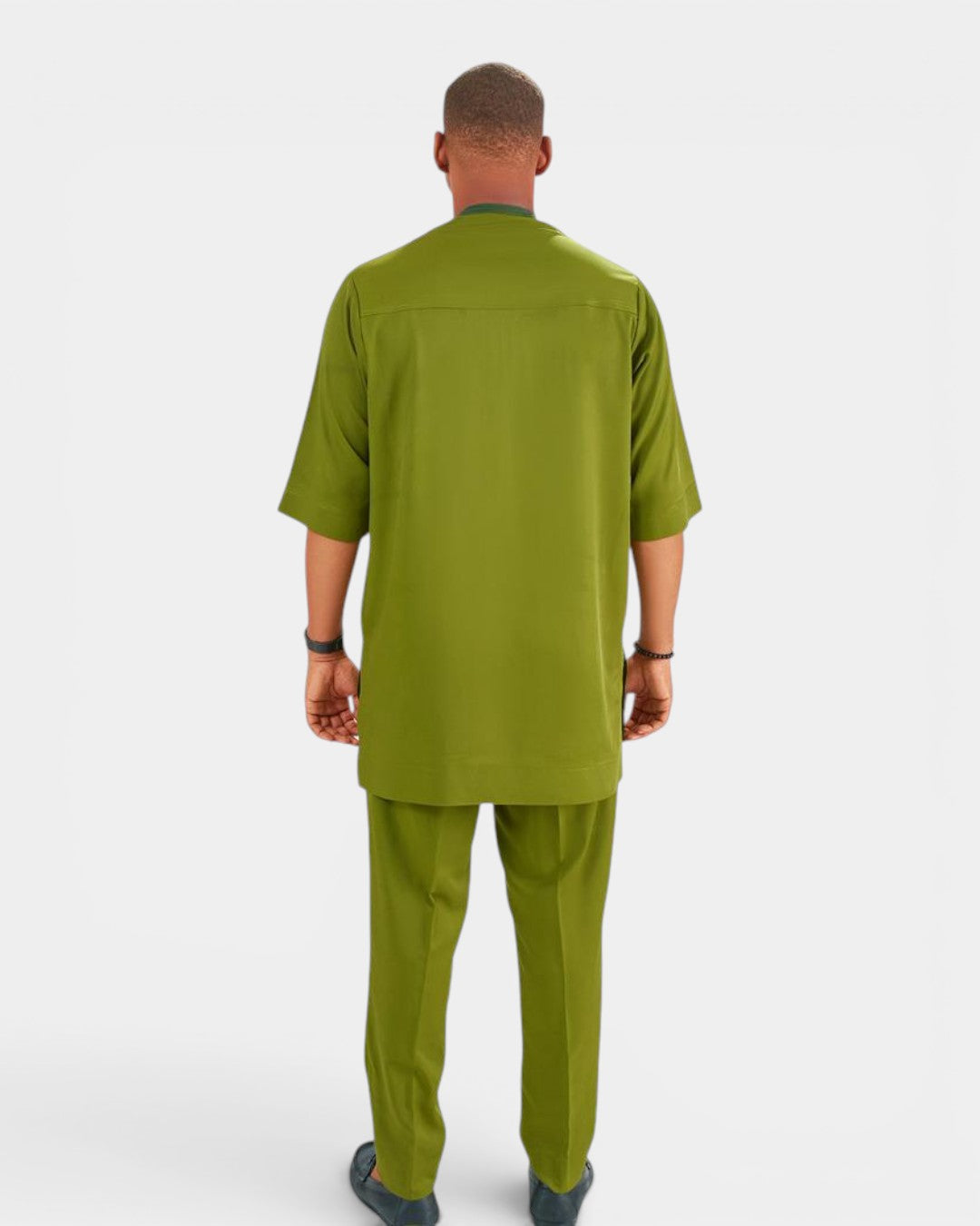 A man viewed from the back wearing an olive green short-sleeve scrub top and matching scrub pants against a light gray studio background.